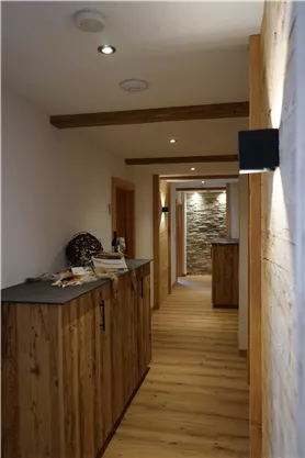 A rustic hallway with wooden paneling and natural light. On the left side, there are several cabinets, and at the end of the hallway, a stone wall is visible.