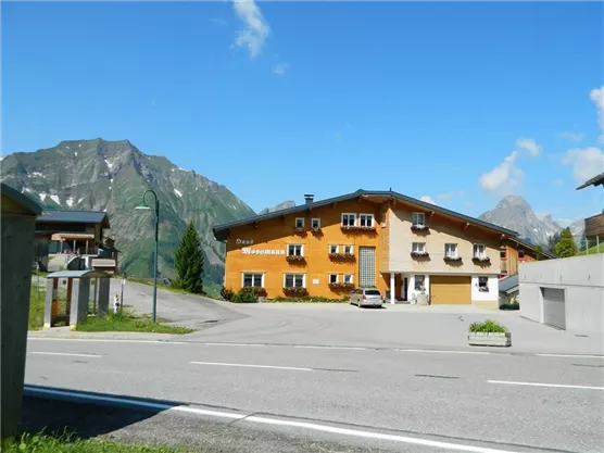 Ein gemütliches Gebäude in einer Berglandschaft mit blauem Himmel. Im Vordergrund ist eine Straße und einige Autos zu sehen.