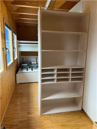 A modern room with wood paneling and large windows. On the left side is a white bookshelf, and in the background is a loft bed.
