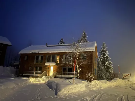 A cozy house in the snow at night, illuminated by a warm light. Surrounded by snow-covered trees and a clear, twilight sky.