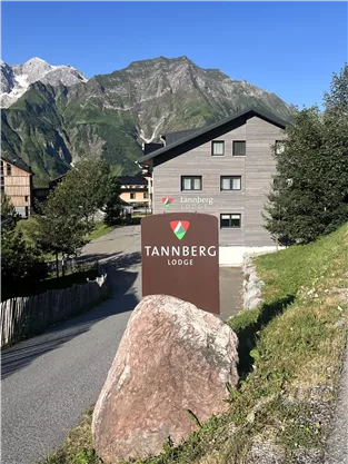 A building with the sign "Tannberg Lodge" is located in a picturesque mountain landscape. In the background, tall mountains and a clear sky can be seen.