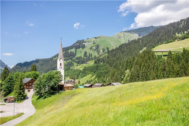 Eine malerische Landschaft mit grünen Wiesen und Bergen im Hintergrund. Im Vordergrund steht eine Kirche mit spitzem Turm, umgeben von traditionellen Chalets.