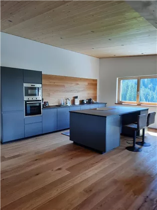A modern kitchen with a large island block and hardwood flooring. Large windows provide plenty of natural light and a view of the surroundings.