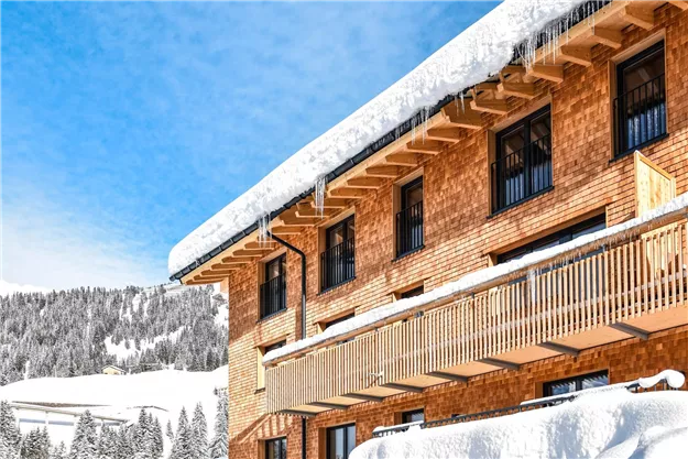 Ein rustikales Gebäude in den Bergen, umgeben von Schnee. Der Himmel ist klar und blau, und das Dach ist mit einer dickeren Schneeschicht bedeckt.