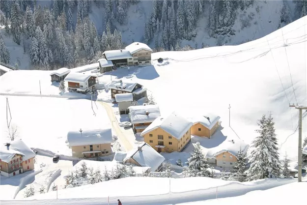 Eine verschneite Landschaft mit kleinen, bunten Häusern. Umgeben von hohen, schneebedeckten Bäumen und einer ruhigen Winteratmosphäre.