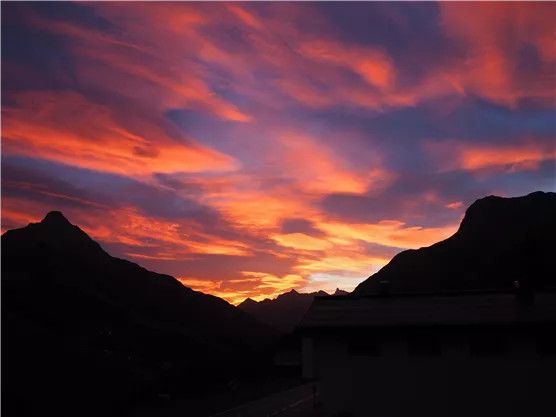A breathtaking sunset over mountains with glowing, colorful clouds. The silhouettes of the mountains and a building are visible in the foreground.