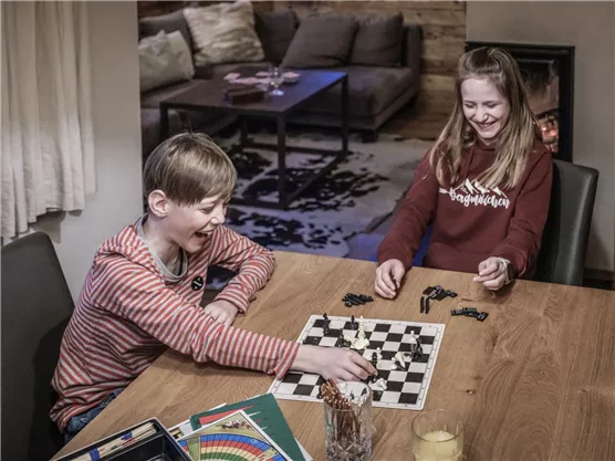 Zwei Kinder spielen Schach an einem Holztisch in einem gemütlichen Raum. Sie schauen sich gegenseitig an und haben Spaß während des Spiels.