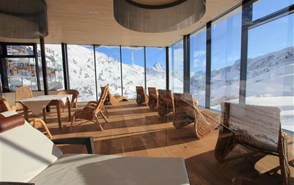 A modern restaurant with large windows and a view of snow-covered mountains. Cozy wooden and wicker furniture creates an inviting atmosphere.