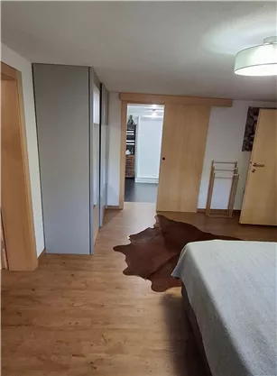 Ein modernes Zimmer mit Holzfußboden und neutralen Farben. Im Vordergrund liegt ein Teppich und im Hintergrund sind zwei Türen sichtbar.