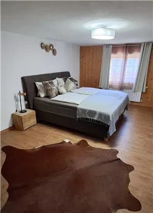 Ein modernes Schlafzimmer mit einem großen Bett und dekorativen Kissen. Der Boden ist mit einem Teppich aus Tierhaut bedeckt und das Zimmer hat viel Tageslicht.