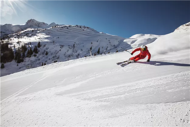 Ein Skifahrer in einem roten Anzug fährt eine schneebedeckte Piste hinunter. Die Umgebung ist von Bergen und klarem, blauem Himmel umgeben.