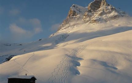 Eine verschneite Berglandschaft mit einem kleinen Holzhaus im Vordergrund. Im Hintergrund ragt ein imposanter Gipfel in einen klaren Himmel.