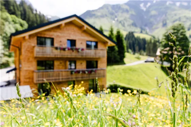 Ein charmantes Holzhaus inmitten einer grünen Landschaft. Wiesenblumen blühen im Vordergrund und die Berge sind im Hintergrund sichtbar.