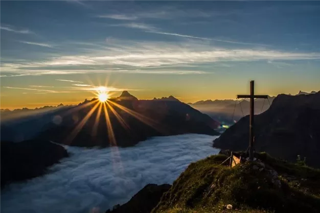 A breathtaking sunrise over the mountains with a cross silhouette in the foreground. Dense clouds spread out in the valley as the sun rises brilliantly.