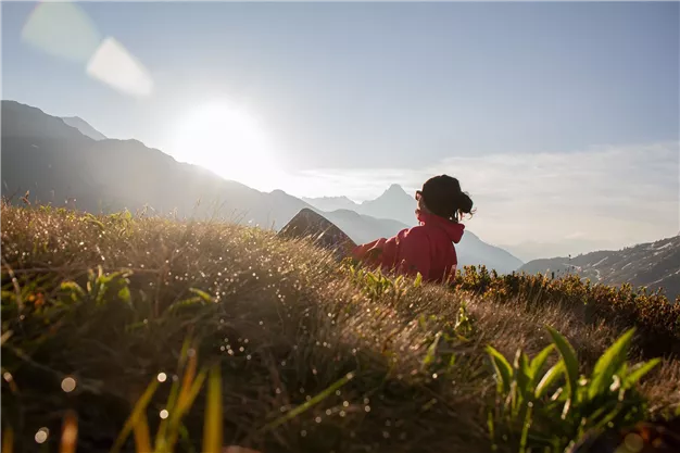 A person is sitting on the grass and looking at the mountains. The sun is shining in the background, and the sky is clear.