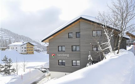 A modern chalet in the snow with a clear facade. In the background, snowy hills and other buildings can be seen.