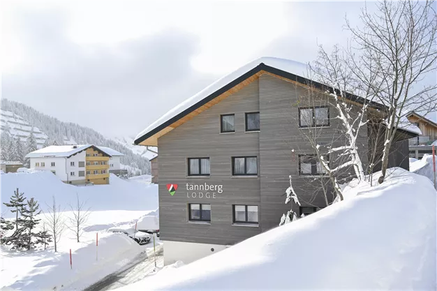 A modern chalet in the snow with a clear facade. In the background, snowy hills and other buildings can be seen.
