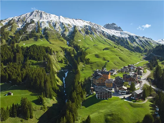 A picturesque mountain landscape with snow-covered peaks and lush green meadows. In the foreground lies a charming village with buildings and a clear blue sky above.