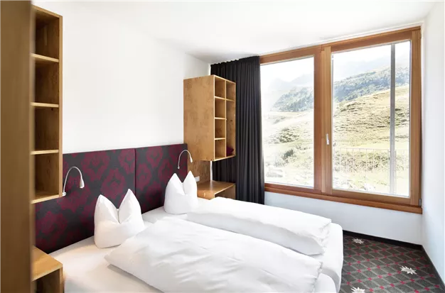Ein modernes Schlafzimmer mit zwei Betten und einem großen Fenster, das einen Blick auf die Berge bietet. Die Einrichtung ist schlicht und elegant, mit Holzregalen und einer gemütlichen Atmosphäre.