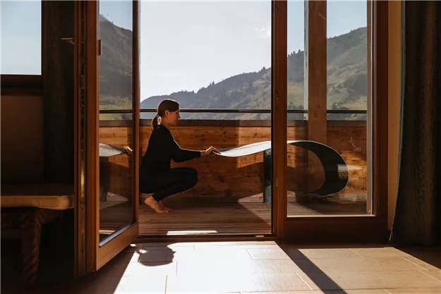 A person is sitting on the balcony and looking at the view of the mountains. The room is brightly lit and has large windows.