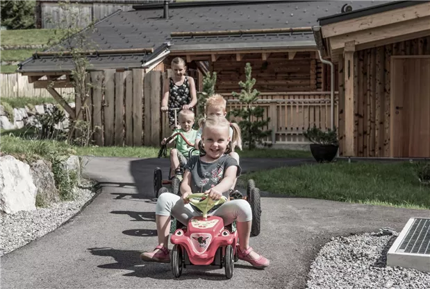 Kinder fahren auf einem Weg mit kleinen Fahrzeugen. Im Hintergrund sind Holzhäuser und Bäume zu sehen.