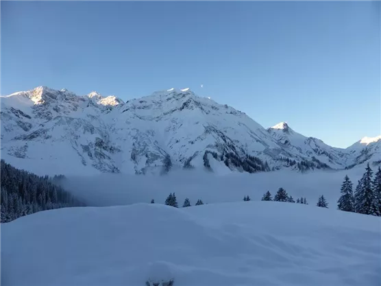 A wintry mountain landscape with snow-covered peaks and a clear sky. In the foreground lies a gentle, snow-covered area.