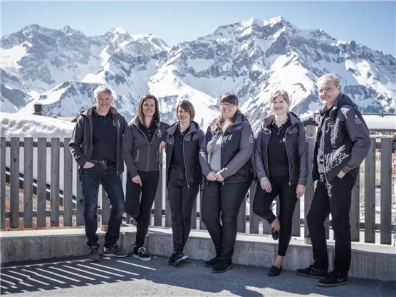 Eine Gruppe von sechs Personen steht vor einer beeindruckenden Berglandschaft mit schneebedeckten Gipfeln. Sie tragen ähnliche Arbeitskleidung und lächeln in die Kamera.
