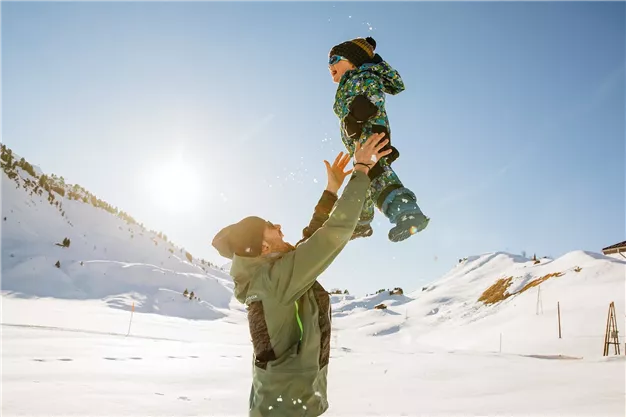 Ein Vater hebt sein Kind hoch in die Luft, umgeben von schneebedeckten Bergen. Die Sonne scheint hell am blauen Himmel.
