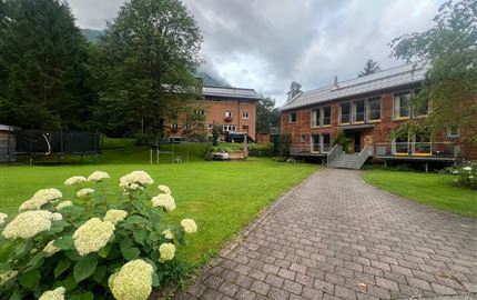 Eine ruhige Gartenlandschaft mit grünen Wiesen und blühenden Sträuchern. Im Hintergrund sind mehrere Gebäude zu sehen, die von Bäumen umgeben sind.