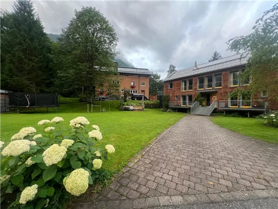 Eine ruhige Gartenlandschaft mit grünen Wiesen und blühenden Sträuchern. Im Hintergrund sind mehrere Gebäude zu sehen, die von Bäumen umgeben sind.