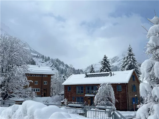 A snowy landscape with two-story buildings and snow-covered trees. The sky is overcast, creating a winter atmosphere.