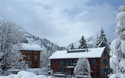 A snowy landscape with two-story buildings and snow-covered trees. The sky is overcast, creating a winter atmosphere.
