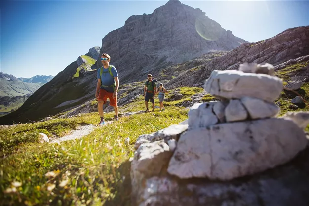 Drei Wanderer erkunden eine malerische Berglandschaft mit grünen Wiesen und hohen Gipfeln im Hintergrund. Ein Steinhaufen befindet sich im Vordergrund, was den Wanderweg markiert.