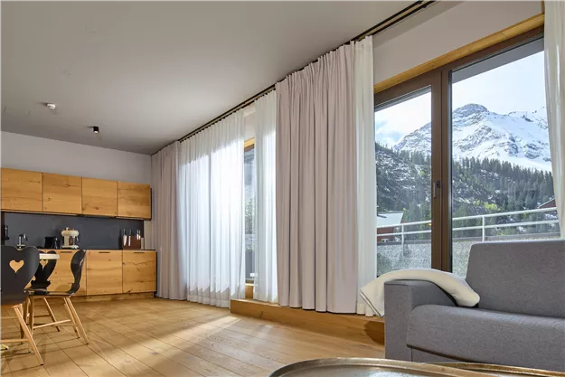 A modern living room with large windows and a view of snow-covered mountains. The room is bright and stylishly furnished with wooden furniture and cozy curtains.