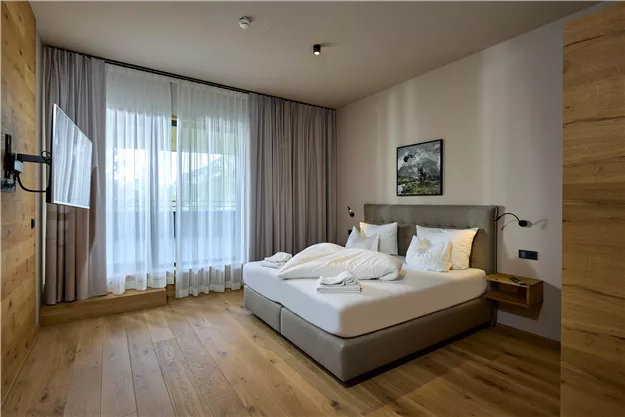 A modern bedroom with a double bed and wooden floor. Large windows provide plenty of daylight and a cozy atmosphere.