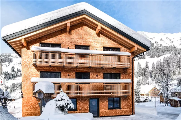 Ein charmantes Holzhaus im Winter, umgeben von schneebedeckten Bäumen. Die Fassade zeigt Balkone und einladende Fenster.