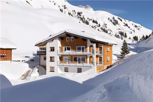 Ein schönes, schneebedecktes Alpenhaus mit Holz- und Glasdetails. Im Hintergrund sind schneebedeckte Berge zu sehen.