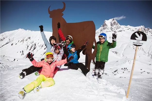 A group of happy children stands in the snow and celebrates. In the background, snow-covered mountains and a noticeable cow sign can be seen.