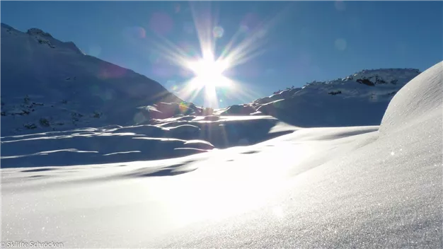 A snowy landscape with high mountains in the background. The sun shines brightly in the sky.