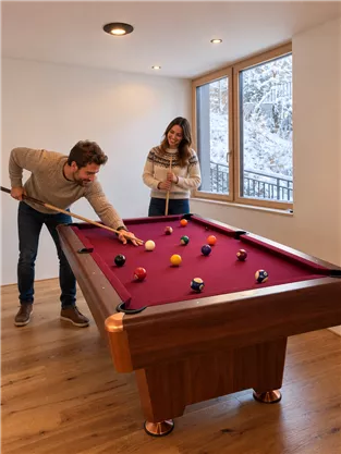 A couple is playing billiards in a cozy room. In the background, you can see snow-covered trees through the windows.