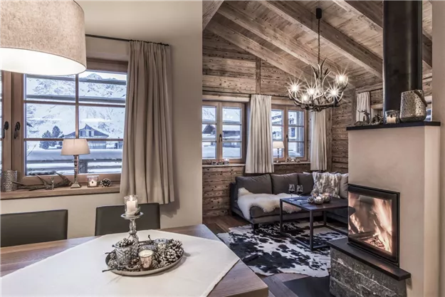 Ein gemütliches Wohnzimmer mit Holzdecke und großen Fenstern. Der Kamin sorgt für eine warme Atmosphäre in der winterlichen Umgebung.