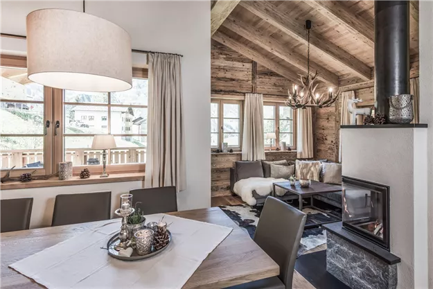 A cozy living room with wooden paneling and a large window front. The room is modernly furnished and has an open fireplace.