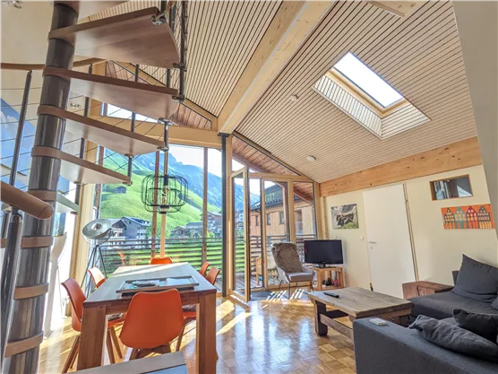 A modern living room with wooden elements and large windows. In the background are green mountains and a clear sky.