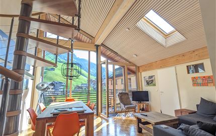 Ein modernes Wohnzimmer mit Holzelementen und großen Fenstern. Im Hintergrund sind grüne Berge und ein klarer Himmel zu sehen.