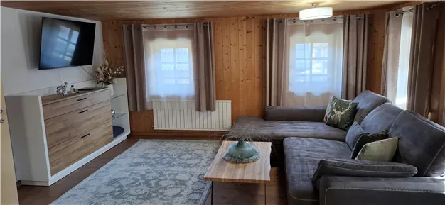 Ein gemütliches Wohnzimmer mit einer grauen Couch und einem Holztisch. Im Raum befinden sich ein Fernseher und große Fenster mit Vorhängen.
