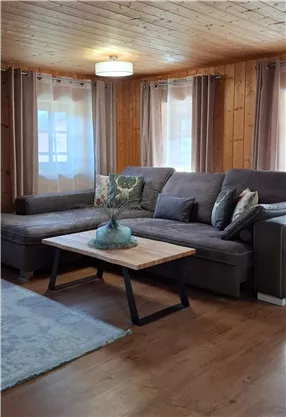 Ein gemütliches Wohnzimmer mit einem grauen Sofa und einem Holztisch. Fenster mit Vorhängen sorgen für eine helle, einladende Atmosphäre.