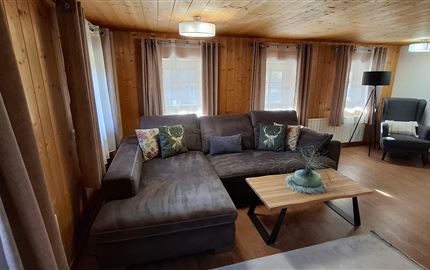 Ein gemütliches Wohnzimmer mit einem großen grauen Sofa und einem Holzkaffee Tisch. Helle Fenster sorgen für eine freundliche Atmosphäre.