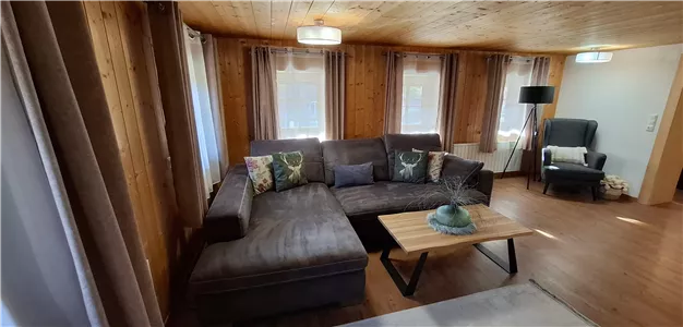 Ein gemütliches Wohnzimmer mit einem großen grauen Sofa und einem Holzkaffee Tisch. Helle Fenster sorgen für eine freundliche Atmosphäre.